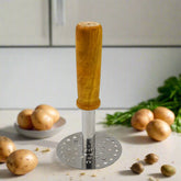 A stainless steel potato masher with a light brown wooden handle stands upright on a kitchen counter with raw potatoes