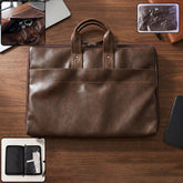A brown faux leather laptop bag with handles and a front pocket, displayed on a wooden surface with a laptop and phone
