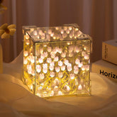 A clear acrylic cube lamp illuminates a cluster of white tulip-shaped lights on a light fabric surface.