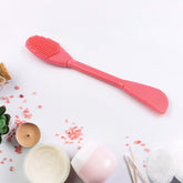A pink dual-ended silicone tool with a textured brush on one end and a spatula on the other, surrounded by skincare items.