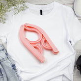 A pink plastic folding hanger rests on a white t-shirt, surrounded by light-wash denim and white flowers.