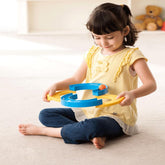 A young girl in a yellow shirt and blue pants sits on the floor, holding a blue and yellow figure-eight track with an orange