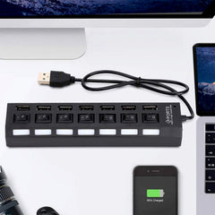A black 7-port USB hub with white LED indicators is positioned on a desk with a laptop, monitor, and smartphone.