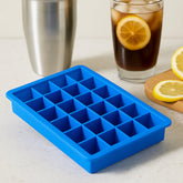 A blue silicone ice cube tray with 24 square cavities sits on a white counter next to a drink and shaker.