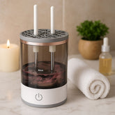 A white and clear makeup brush cleaner with two white brush holders, filled with swirling purple liquid, sits on a counter