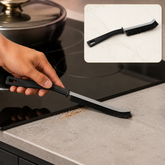 A hand uses a black and gray crevice cleaning brush to sweep crumbs off a kitchen counter next to a stovetop.