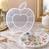 A clear apple-shaped organizer box with a heart-shaped center, filled with various colorful jewelry on a marble surface.
