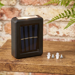 A black solar panel light with mounting screws and anchors on a wooden surface, with a brick wall background.
