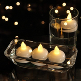 Three white LED floating candles in a clear rectangular dish and one in a tall glass, all lit.