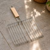 A silver metal grilling basket with a light wooden handle rests on a grey tiled floor, illuminated by sunlight.