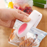 A hand uses a white and pink mini heat sealer to seal a clear bag of snacks on a wooden surface.