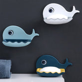 Three whale-shaped soap dishes in white, light blue, and dark blue, each holding a white bar of soap, mounted on a dark gray