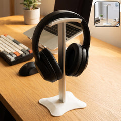 White metal headphone stand on a light wooden desk next to a green plant and stacked books.