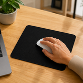 A hand uses a white mouse on a black mouse pad placed on a wooden desk next to a laptop and plant.