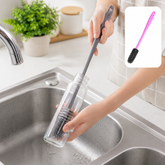 A person cleans a clear glass bottle with a grey silicone brush in a stainless steel sink. A small inset shows a pink