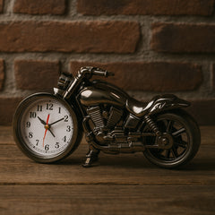 A dark silver motorcycle clock with a white face is displayed on a rustic wooden table against a brick wall.