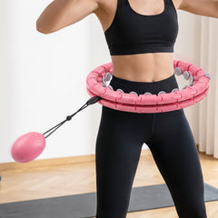A woman in a black sports bra and leggings uses a pink weighted hula hoop with a swinging massage ball.