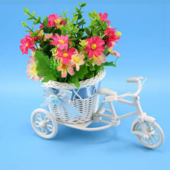 White plastic tricycle planter filled with vibrant pink and yellow artificial flowers and green leaves.