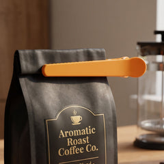 An orange plastic scoop and clip is shown attached to a black coffee bag, sealing it closed.