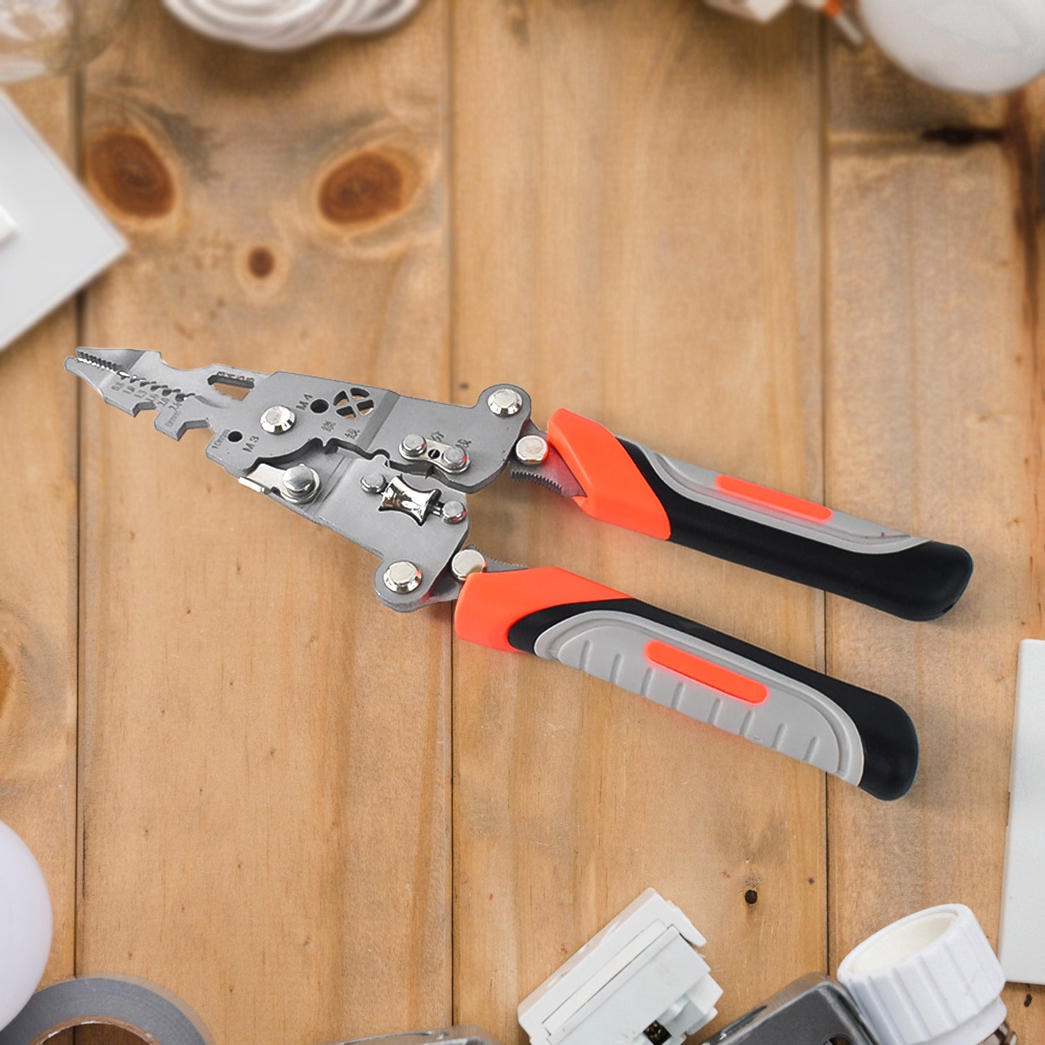 A silver and orange wire stripper tool with black and grey handles rests on a wooden surface surrounded by various