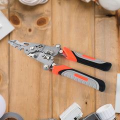 A silver and orange wire stripper tool with black and grey handles rests on a wooden surface surrounded by various