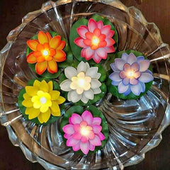 Six glowing lotus flower candles in different colors, floating in a clear, decorative glass bowl filled with water.