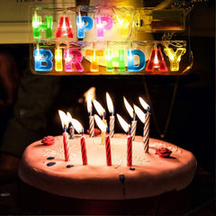 Glowing "Happy Birthday" string lights above a pink frosted cake with lit striped candles.