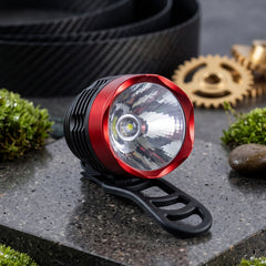 A red and black bicycle headlight with a clear lens sits on a grey stone surface with moss and gears.