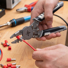 A person's hands operating a gray and black wire crimping tool with a red wire and connector, on a wooden surface.