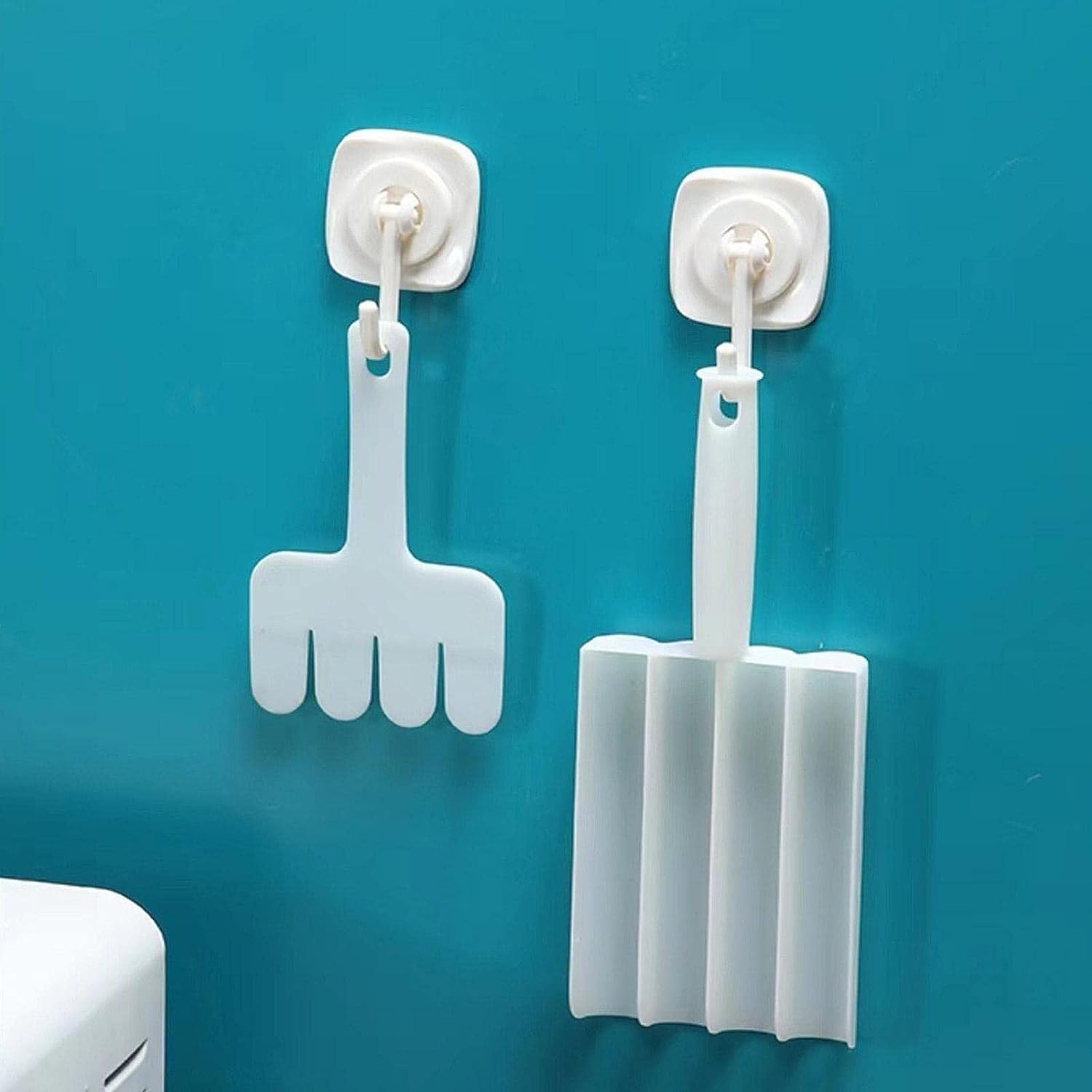 Two white plastic kitchen tools, a four-pronged rake and a four-chambered scoop, hang on wall hooks.