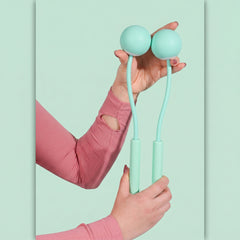 Hands hold the mint green handles and weighted balls of a cordless jump rope against a light green background.