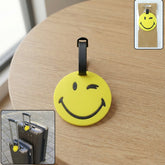 A yellow winking emoji luggage tag with a black strap sits on a wooden table, with insets showing its packaging and use on