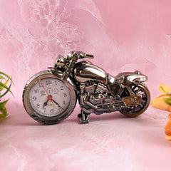 A metallic motorcycle clock with a white face and red hands rests on a pink marble surface.