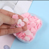 A hand holds pink and white heart and circle shaped paper soap flakes above a clear apple-shaped container filled with more