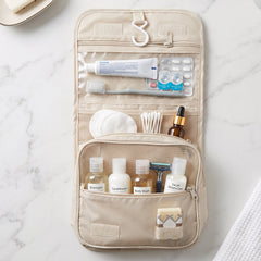 The beige hanging toiletry bag is open, revealing multiple pockets filled with travel essentials like shampoo and a razor.