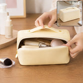 A person's hands open a cream-colored cosmetic bag, revealing various makeup items inside, on a wooden surface.
