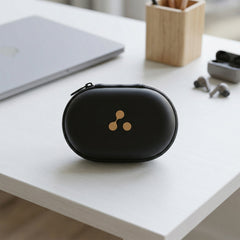 A closed black oval earbud case with a gold three-dot logo sits on a white table next to a laptop.