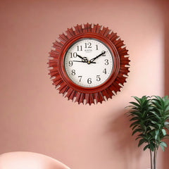 Red sunburst wall clock hanging on a pink wall, with a green potted plant visible on the right side.