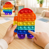 A child's hands gently press the colorful silicone bubbles of an Among Us shaped fidget toy on a wooden table.