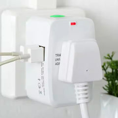 A white universal travel adapter with a green button and red light has a white cable plugged into its side.