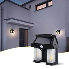 A black solar wall lamp with two clear light panels illuminates a modern grey house exterior at dusk.
