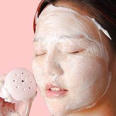 A woman with a foamy face mask uses a pink octopus silicone facial cleanser to gently massage her cheek.