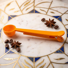 An orange plastic coffee scoop with an integrated bag clip rests on a decorative white and blue marble surface, surrounded