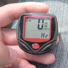 A person's hand holds the small black and red bicycle computer, showing a digital display with numbers.
