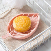 A pink leaf-shaped soap dish with a yellow bar of soap on a beige towel inside a white wire basket.