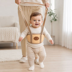 A smiling baby in a cream outfit wears a beige walking harness, held by an adult in a well-lit room.