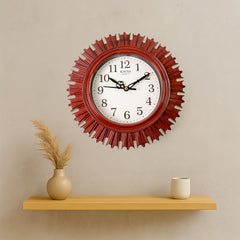 Red sunburst wall clock on a light grey wall, centered above a wooden shelf with a vase and cup.