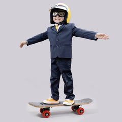 A young boy in a suit, yellow helmet, and goggles stands on a skateboard with red wheels, arms outstretched.