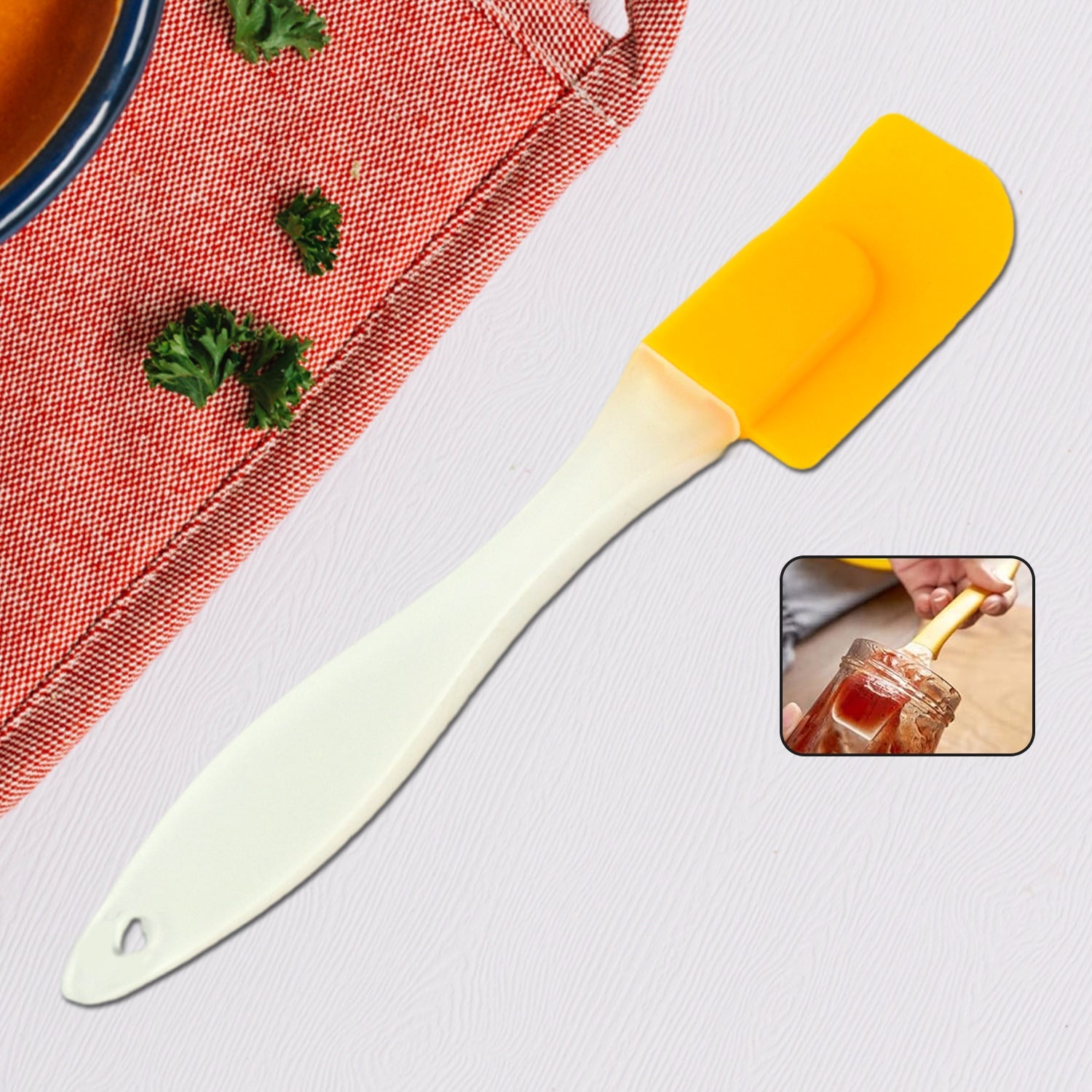 A yellow silicone spatula with a cream handle lies on a grey surface, next to a red fabric and a small inset of a hand using