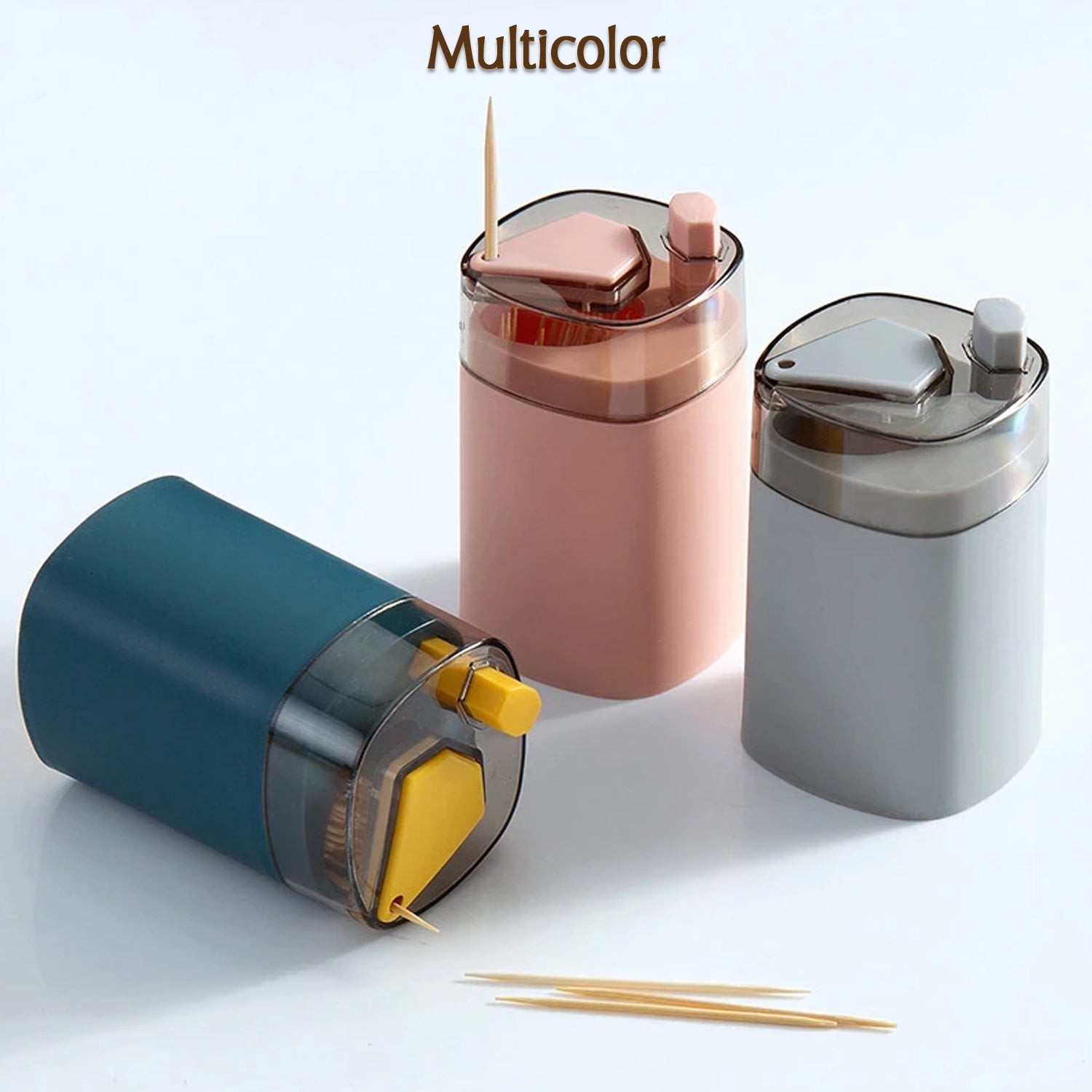 Three automatic toothpick dispensers in teal, pink, and grey, arranged on a white surface with two toothpicks in the